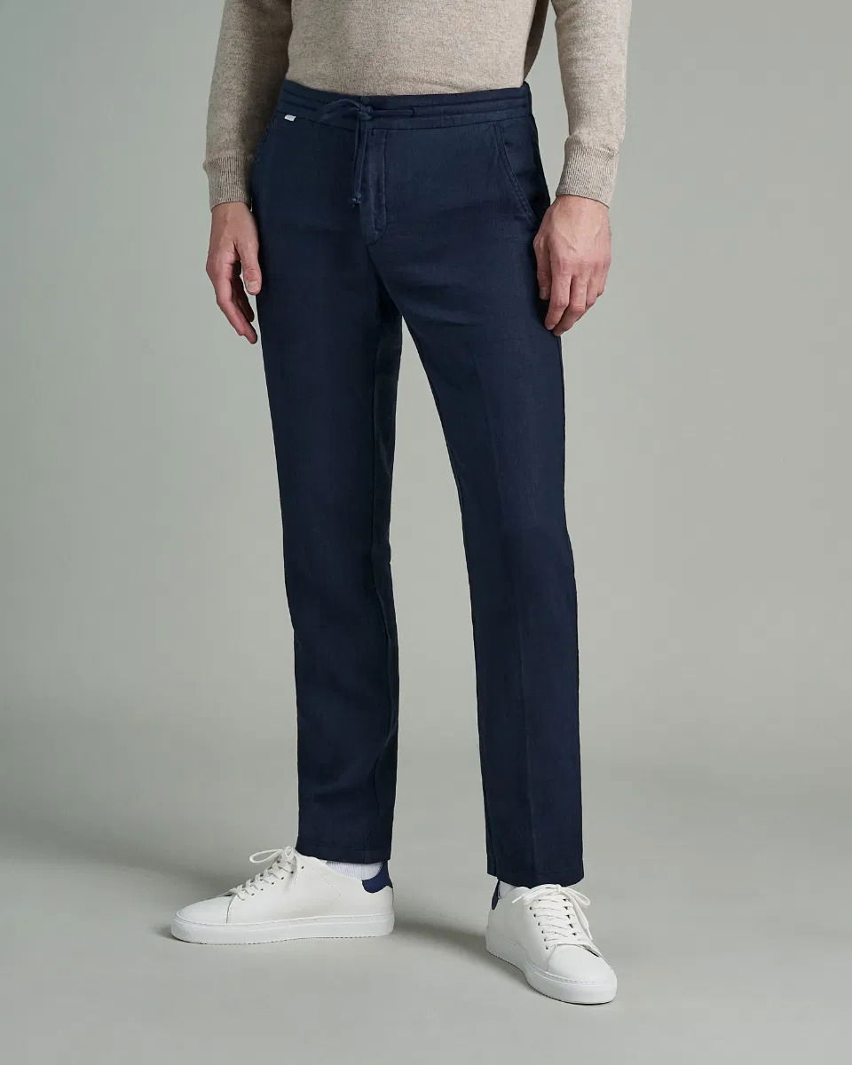 Herren trägt dunkelblaue Joggpants mit Kordel, beigefarbenen Pullover und weiße Sneaker mit blauen Details vor grauem Hintergrund. Herren trägt dunkelblaue Joggpants mit Kordel, beigefarbenen Pullover und weiße Sneaker mit blauen Details vor grauem Hintergrund.