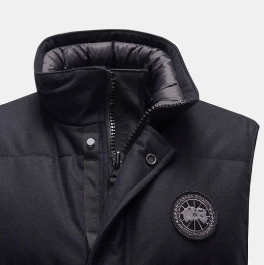 Anschnitt einer schwarzen Herren-Outdoorweste mit hohem Kragen, Reißverschluss, Druckknöpfen und rundem Logo auf der Brust.