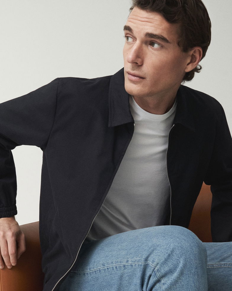 Mann sitzt, trägt dunkelblauen Blouson, weißes T-Shirt und hellblaue Jeans, schaut nach rechts, heller Hintergrund.