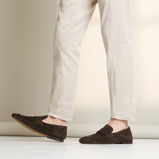 Beine in beigefarbenen Herrenhosen mit dunkelbraunen Wildleder-Loafern, stehend auf hellem Boden vor hellem Hintergrund.