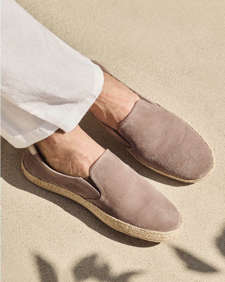 Hellbraune Herren-Espadrilles aus Rauleder mit Jutesohle, getragen zu weißen Hosen, auf hellem Boden. Hellbraune Herren-Espadrilles aus Rauleder mit Jutesohle, getragen zu weißen Hosen, auf hellem Boden.