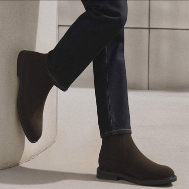 Dunkelbraune Chelsea-Boots aus Leder, getragen zu dunkler Jeans, ein Fuß lehnt an weißer Wand vor grauer Kachelwand.