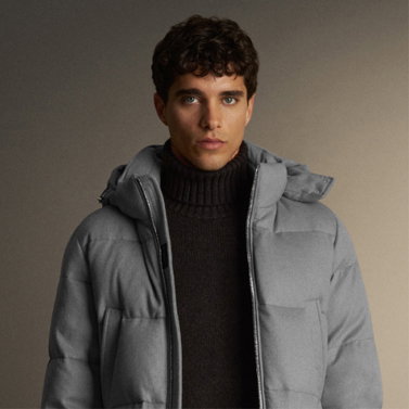 Junger Mann mit lockigem Haar trägt schwarzen Rollkragenpullover und graue Daunenjacke vor neutralem Hintergrund.