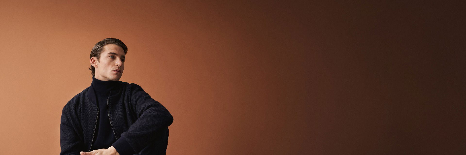 Sitzender Mann mit navyblauem Rollkragenpullover und Blouson vor braunem Hintergrund, nachdenklich nach links schauend. Sitzender Mann mit navyblauem Rollkragenpullover und Blouson vor braunem Hintergrund, nachdenklich nach links schauend.