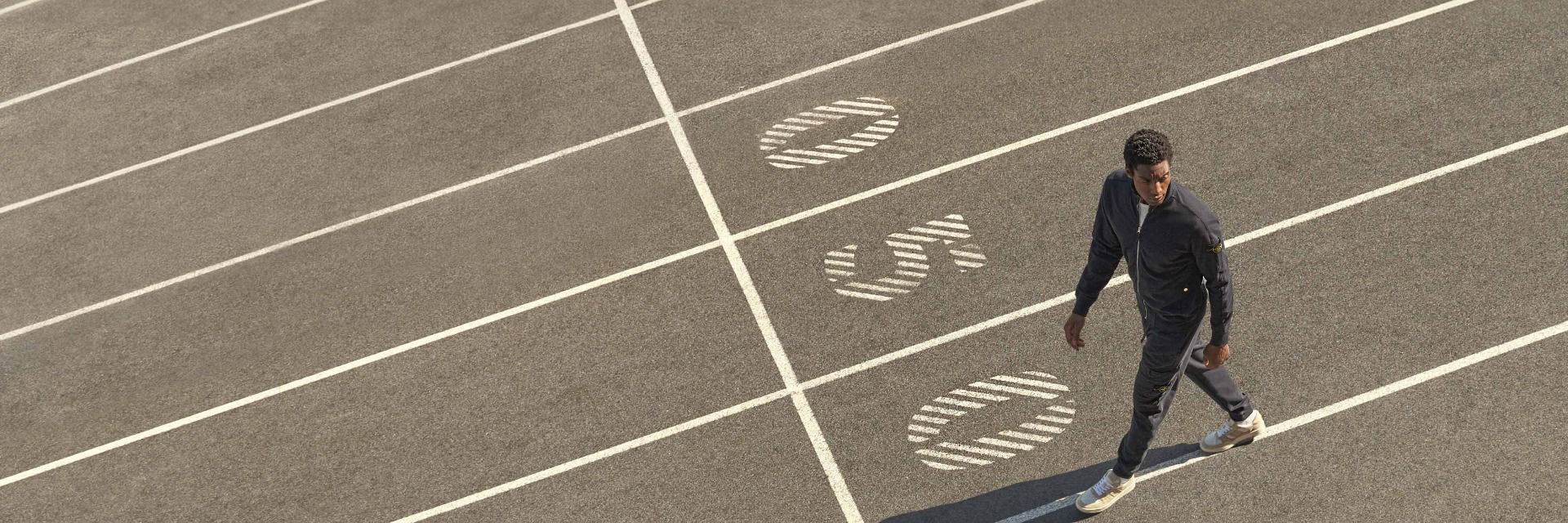 Mann in dunkelgrauem Athleisure-Outfit mit weiß-beigen Sneakern geht auf einer Laufbahn mit weißen Markierungen. Mann in dunkelgrauem Athleisure-Outfit mit weiß-beigen Sneakern geht auf einer Laufbahn mit weißen Markierungen.