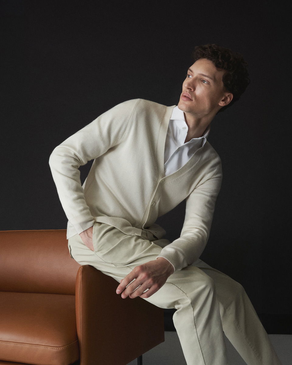 Junger Mann in weißem Hemd, cremefarbenem Cardigan und beiger Hose sitzt auf der Armlehne einer braunen Couch vor dunklem Hintergrund.