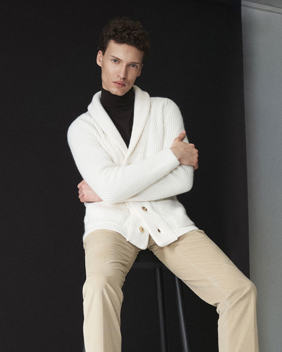 Junger Mann sitzt auf Hocker, trägt weißen Cardigan, schwarzen Rollkragenpullover und beige Cordhose vor dunklem Hintergrund.