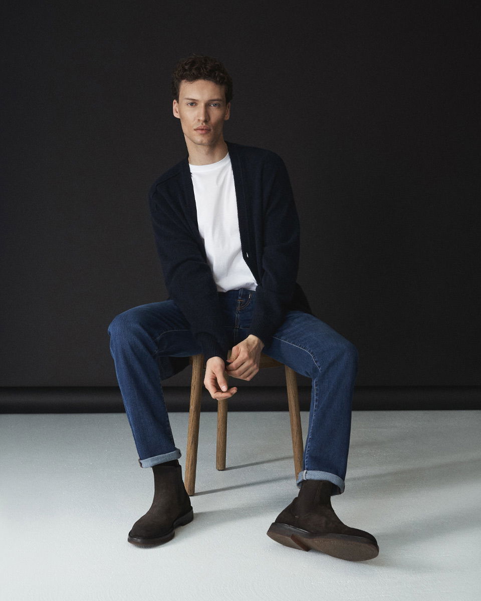 Mann sitzt auf Hocker, trägt navy Cardigan, weißes Shirt, blaue Jeans, braune Chelsea Boots vor schwarzem Hintergrund.