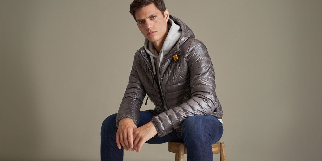 Mann sitzt auf Holz-Hocker, trägt silber-graue Daunenjacke, hellgrauen Hoodie und dunkelblaue Jeans vor beigem Hintergrund. Mann sitzt auf Holz-Hocker, trägt silber-graue Daunenjacke, hellgrauen Hoodie und dunkelblaue Jeans vor beigem Hintergrund.