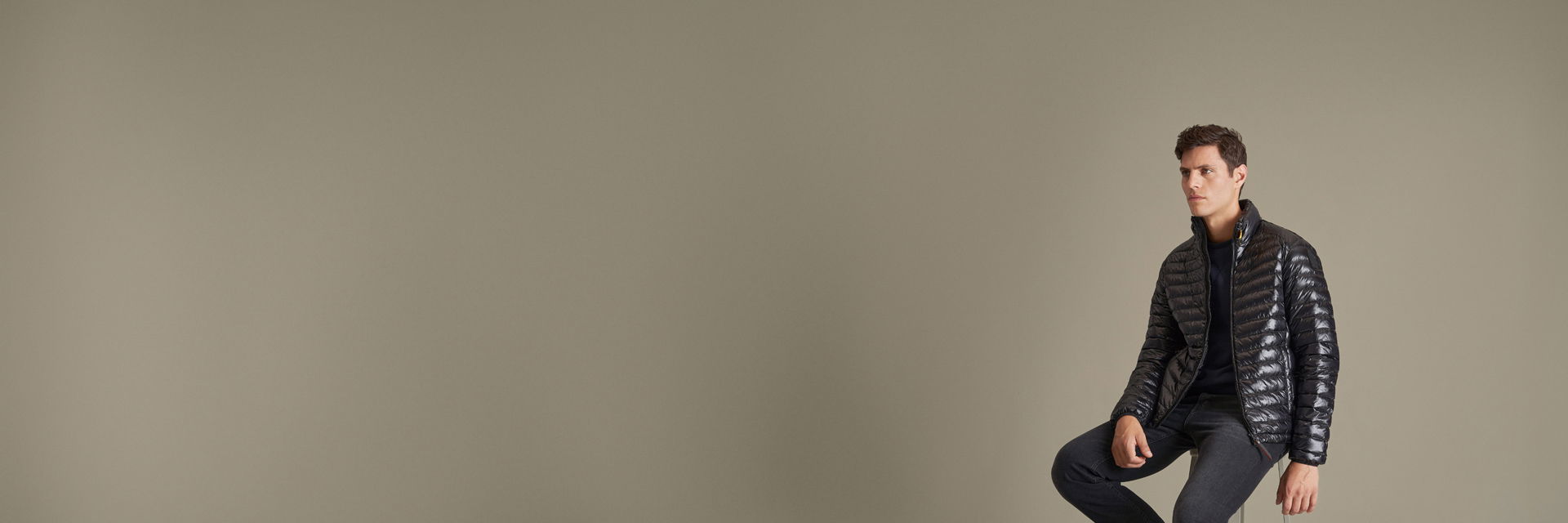 Junger Mann sitzt auf Hocker, trägt schwarze Daunenjacke, schwarzen Pullover und schwarze Jeans vor beige Hintergrund. Junger Mann sitzt auf Hocker, trägt schwarze Daunenjacke, schwarzen Pullover und schwarze Jeans vor beige Hintergrund.