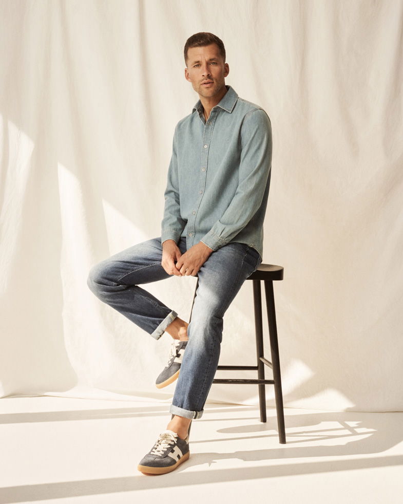 Mann sitzt auf schwarzem Hocker, trägt hellblaues Jeanshemd, rauchblaue Jeanshose und graue Sneaker mit weißen Details.