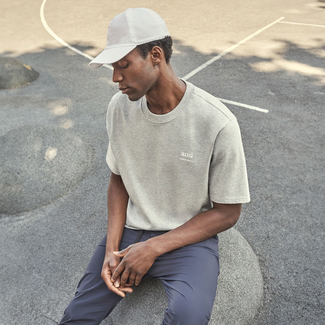 Mann mit weißer Baseball-Cap, grauem Oversized-T-Shirt und blauer Hose sitzt auf rundem Stein auf Asphalt mit weißen Linien. Mann mit weißer Baseball-Cap, grauem Oversized-T-Shirt und blauer Hose sitzt auf rundem Stein auf Asphalt mit weißen Linien.
