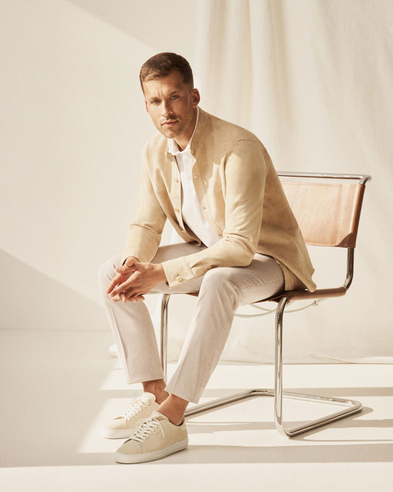 Mann sitzt auf modernem Stuhl, trägt cremefarbene Velourslederjacke, beige Hose, weißes Hemd und cremefarbene Sneaker.