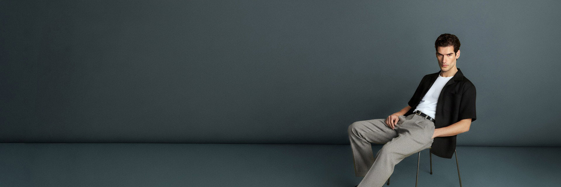 Herr sitzt auf Stuhl, trägt graue Hose, weißen T-Shirt und schwarzes Kurzarmhemd vor schlichtem dunklem Hintergrund. Herr sitzt auf Stuhl, trägt graue Hose, weißen T-Shirt und schwarzes Kurzarmhemd vor schlichtem dunklem Hintergrund.