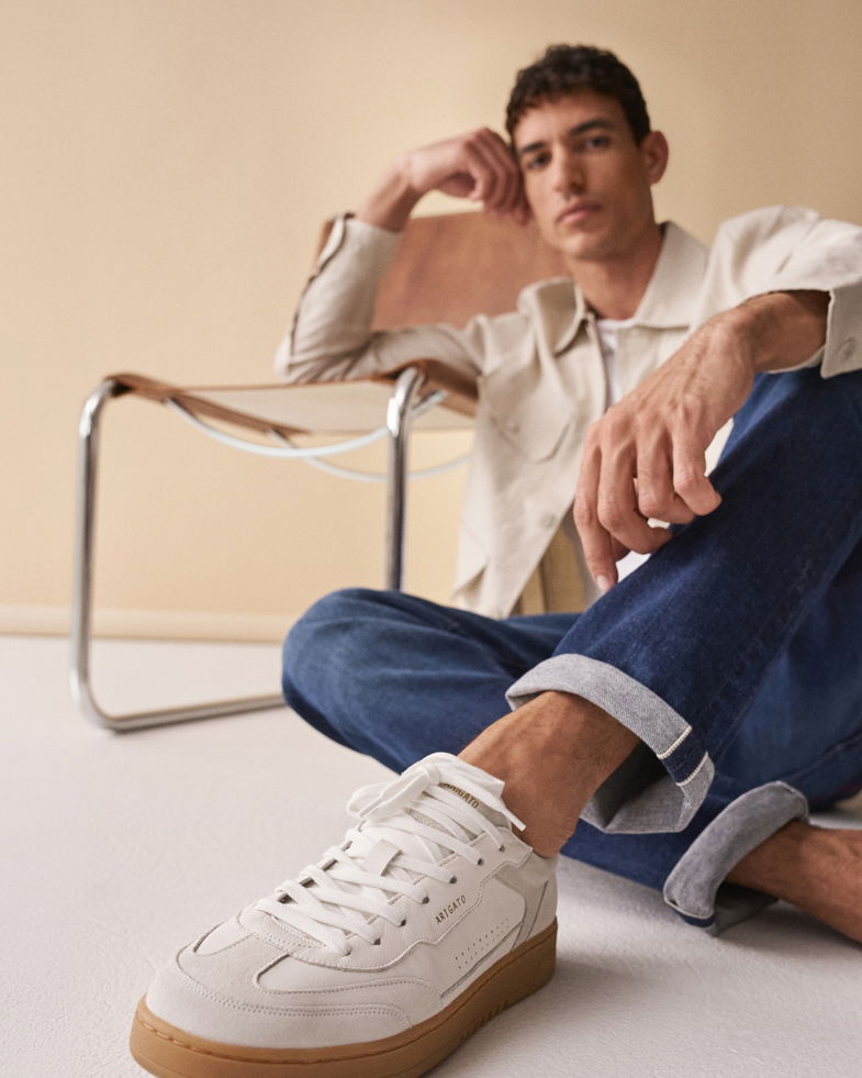 Junger Mann sitzt auf dem Boden, trägt beige Overshirt, blaue Jeans und weiße Sneaker mit brauner Sohle, Holzstuhl im Hintergrund.