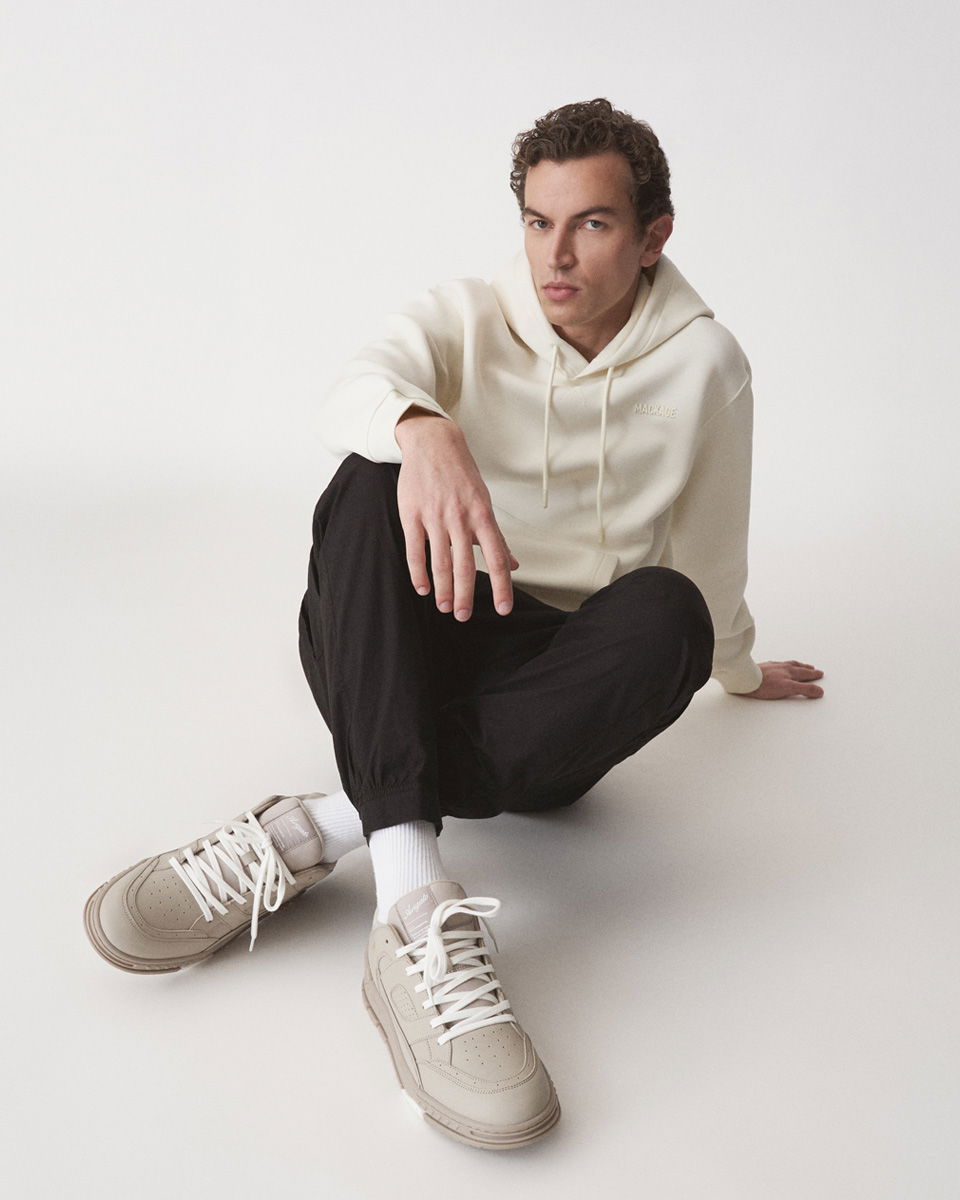 Junger Mann sitzt auf weißem Boden, trägt weißen Hoodie, schwarze Hose, weiße Socken und beige-graue Sneaker. Junger Mann sitzt auf weißem Boden, trägt weißen Hoodie, schwarze Hose, weiße Socken und beige-graue Sneaker.