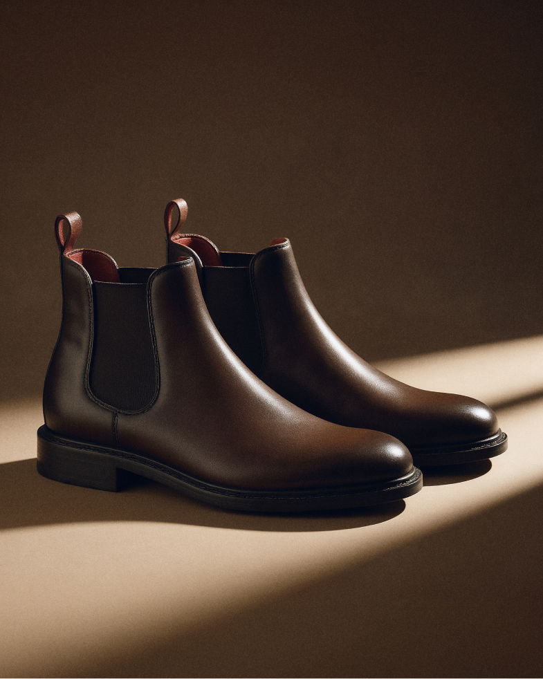 Dunkelbraune Chelsea-Boots aus Glattleder für Herren, stehend auf hellbraunem Untergrund mit seitlichem Elastikeinsatz. Dunkelbraune Chelsea-Boots aus Glattleder für Herren, stehend auf hellbraunem Untergrund mit seitlichem Elastikeinsatz.