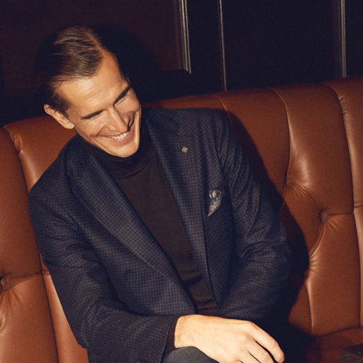 Mann mit dunkelblauem Sakko und schwarzem Rollkragenpullover sitzt lächelnd auf brauner Ledercouch in gemütlicher Umgebung. Mann mit dunkelblauem Sakko und schwarzem Rollkragenpullover sitzt lächelnd auf brauner Ledercouch in gemütlicher Umgebung.