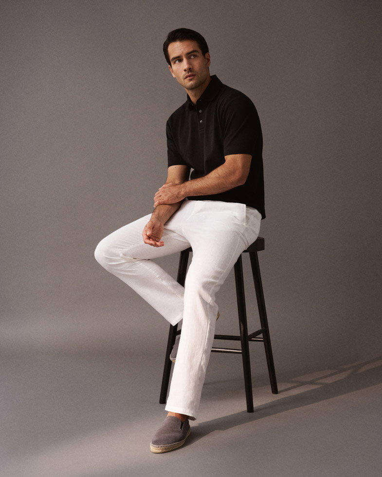 Mann sitzt auf schwarzem Hocker, trägt schwarzes Poloshirt, weiße Hose und hellgraue Espadrilles, schaut nach links. Mann sitzt auf schwarzem Hocker, trägt schwarzes Poloshirt, weiße Hose und hellgraue Espadrilles, schaut nach links.