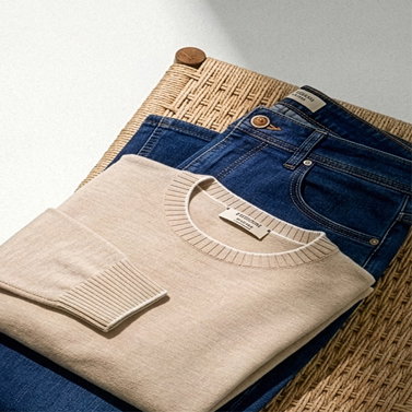 Beiger Rundhalspullover und blaue Jeans ordentlich gefaltet auf einem gewebten Rattanhocker liegend.