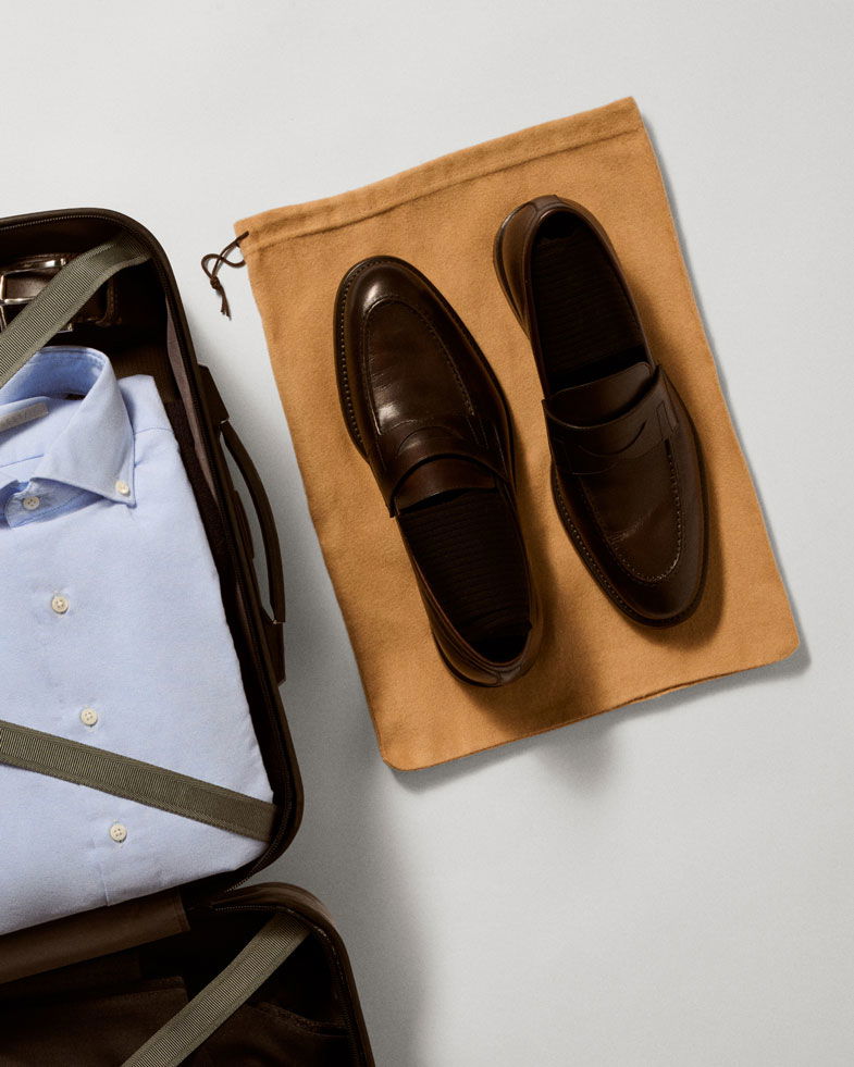 Dunkelbraune Herren-Lederloafer auf beigem Schuhbeutel neben offenem Koffer mit hellblauem Hemd, auf hellem Untergrund.