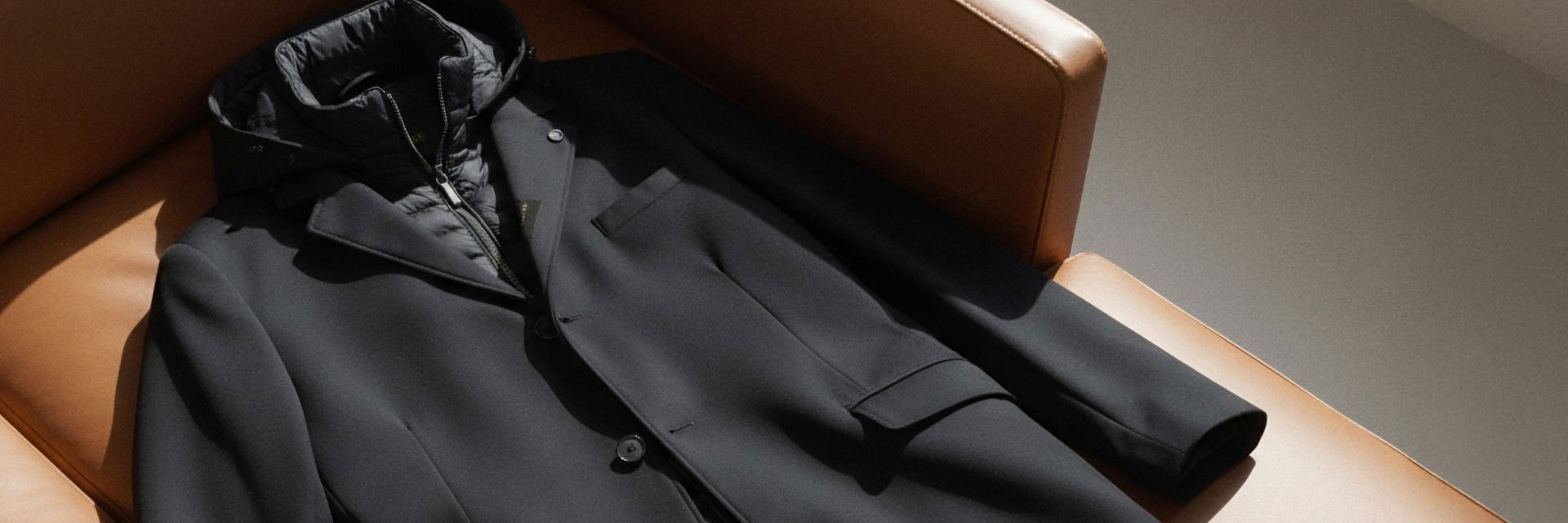 Schwarze Zwei-in-eins-Jacke aus Wollmantel und Daunenweste liegt auf braunem Ledersessel im Sonnenlicht. Schwarze Zwei-in-eins-Jacke aus Wollmantel und Daunenweste liegt auf braunem Ledersessel im Sonnenlicht.