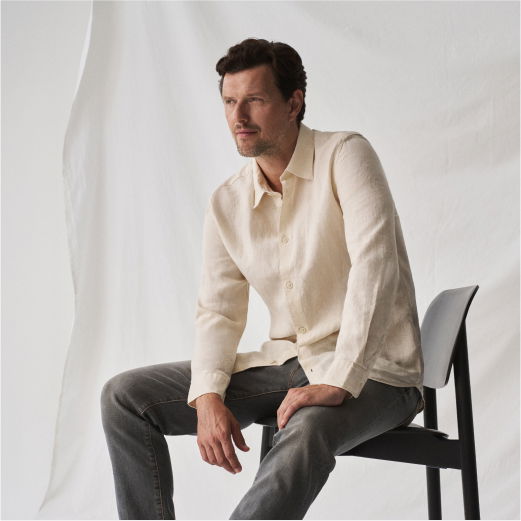 Mann mit cremefarbenem Hemd und grauer Hose sitzt nachdenklich auf schwarzem Stuhl vor weißem Hintergrund im Studio.