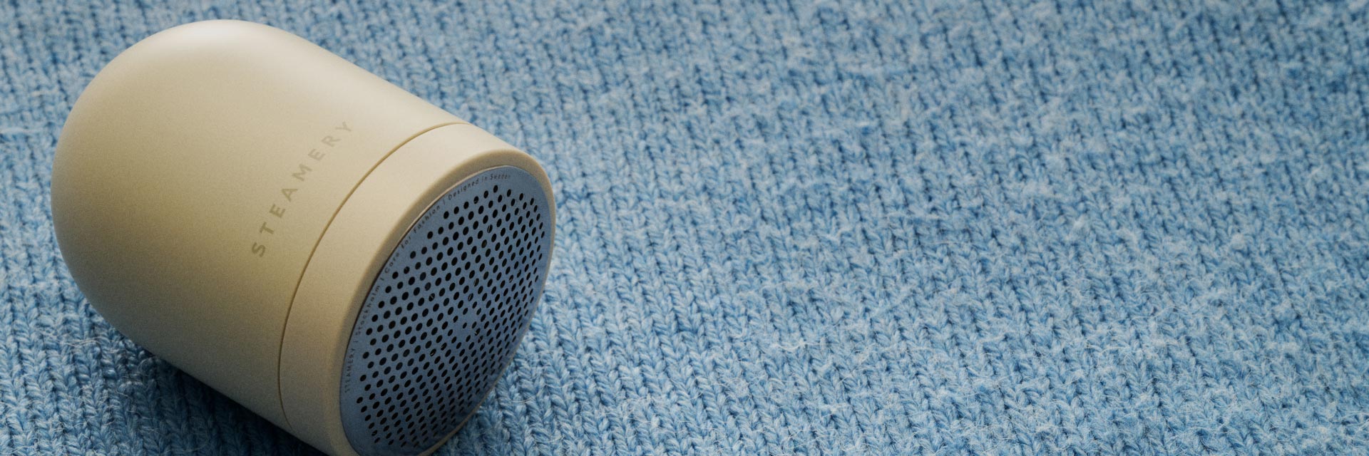 Beiger Fusselrasierer liegt auf hellblauem, grob gestricktem Pullover, Vorderseite mit gelochtem Metallgitter sichtbar. Beiger Fusselrasierer liegt auf hellblauem, grob gestricktem Pullover, Vorderseite mit gelochtem Metallgitter sichtbar.