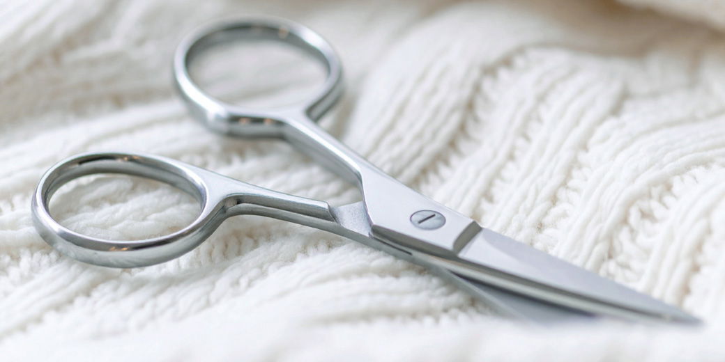 Silberne Nagelschere liegt aufgeklappt auf einem weißen, grob gestrickten Pullover, Nahaufnahme. Silberne Nagelschere liegt aufgeklappt auf einem weißen, grob gestrickten Pullover, Nahaufnahme.