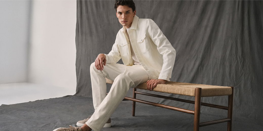 Junger Mann sitzt auf geflochtener Bank, trägt cremefarbenen Denim-Look mit Jacke, Hose und beigen Sneakern vor grauem Hintergrund.