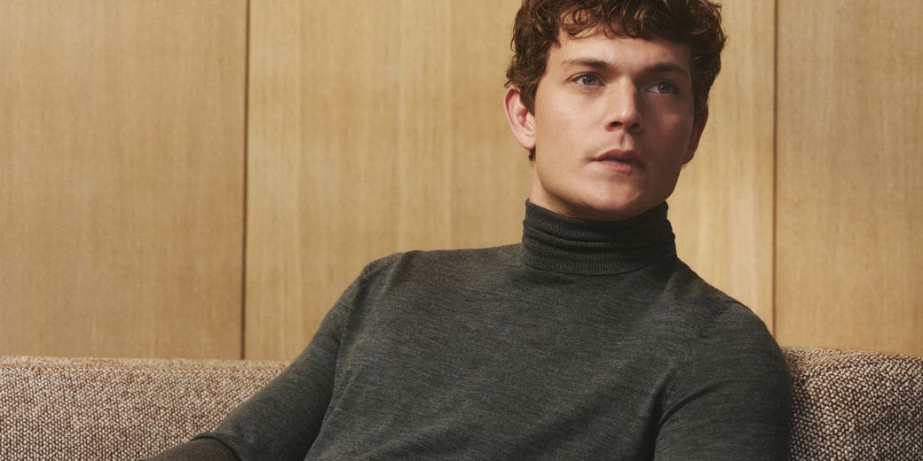 Junger Mann mit lockigem Haar sitzt auf beiger Couch und trägt dunkelgrauen Merino-Rollkragenpullover vor Holzwand.