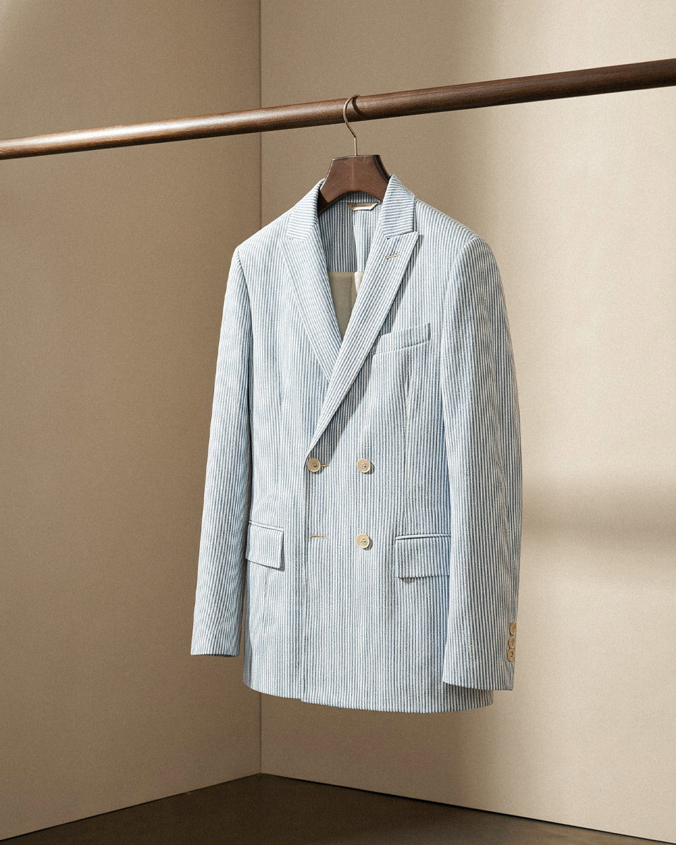 Hellblau-weiß gestreiftes Baumwoll-Sakko mit doppelter Knopfreihe hängt an einer Holzstange vor beiger Wand.