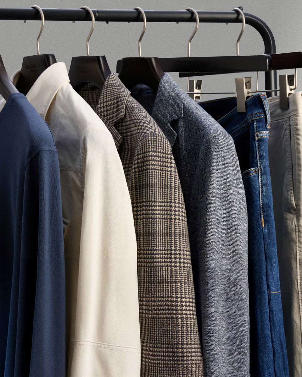 Herrenkleidung auf Kleiderstange, Hemden, Sakkos in Blau, Weiß, Kariert und Jeans sowie hellbeige Hosen aufgehängt.