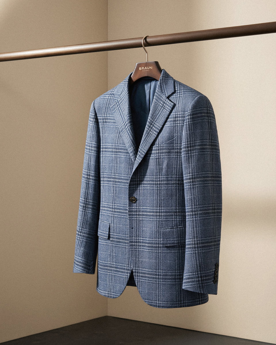 Hellblau-dunkelblau kariertes Wollsakko, einreihig mit zwei Taschen, auf einem Holzkleiderbügel an einer Kleiderstange hängend.