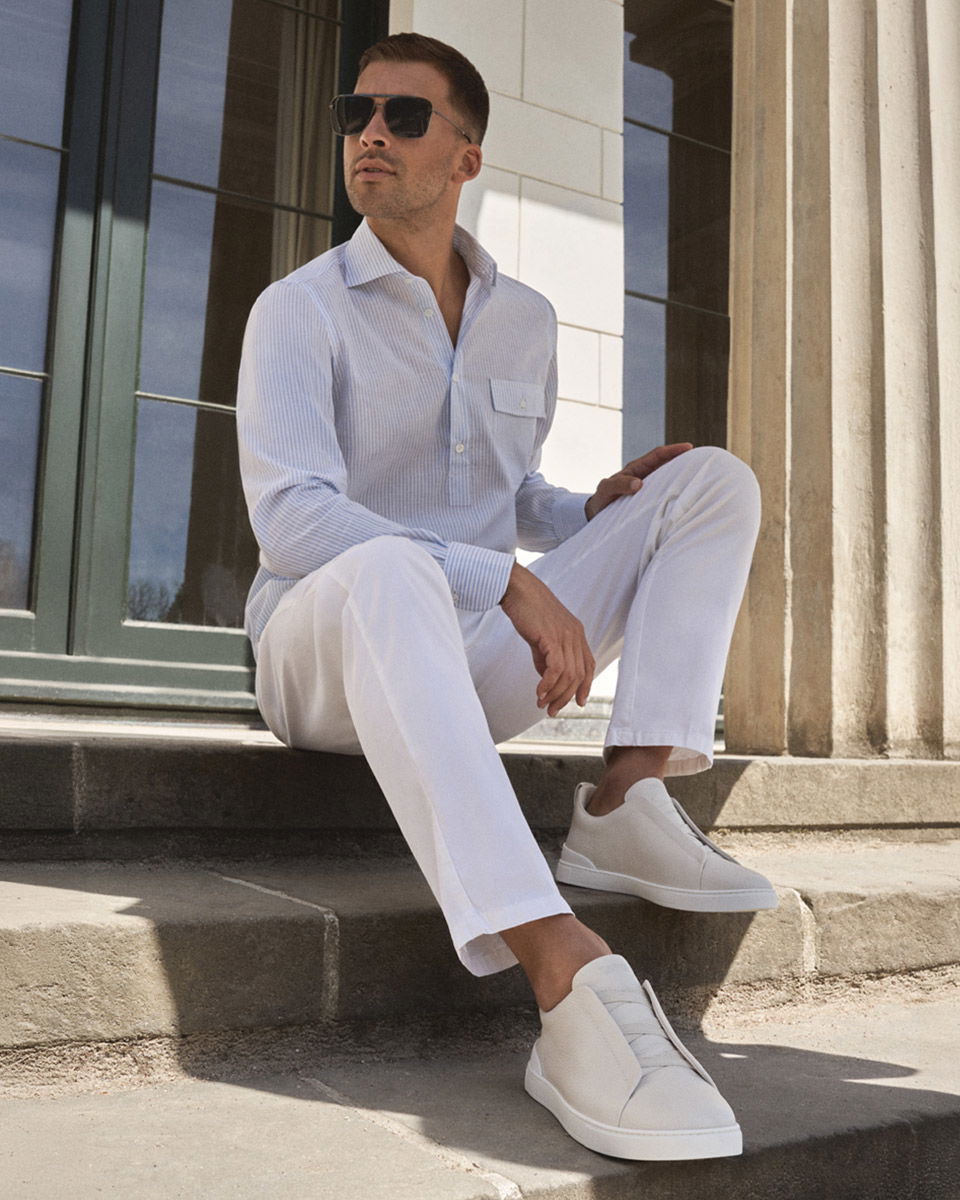 Mann mit weißer Hose, hellblauem Hemd und weißen Sneakern sitzt auf Steintreppe vor Gebäude mit Säulen und Glasfenstern.