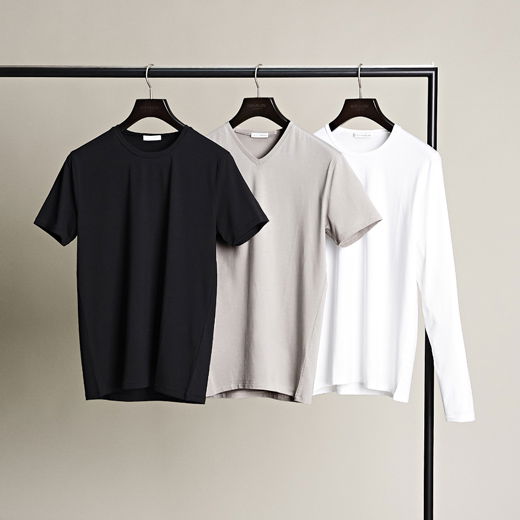 Schwarzes T-Shirt, hellgraues V-Neck T-Shirt und weißes Langarmshirt hängen auf schwarzen Kleiderbügeln an einer Stange vor beige Hintergrund. Schwarzes T-Shirt, hellgraues V-Neck T-Shirt und weißes Langarmshirt hängen auf schwarzen Kleiderbügeln an einer Stange vor beige Hintergrund.