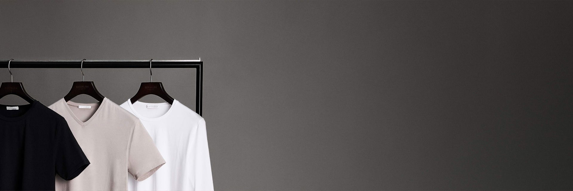 Schlichte T-Shirts in Schwarz, Beige mit V-Ausschnitt und Weiß mit Rundhals, aufgehängt an einem schwarzen Kleiderbügel. Schlichte T-Shirts in Schwarz, Beige mit V-Ausschnitt und Weiß mit Rundhals, aufgehängt an einem schwarzen Kleiderbügel.