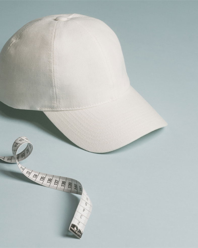 Weiße Baseball-Cap liegt auf hellblauem Untergrund, daneben ein weißes Maßband mit schwarzen Zahlen und Markierungen.