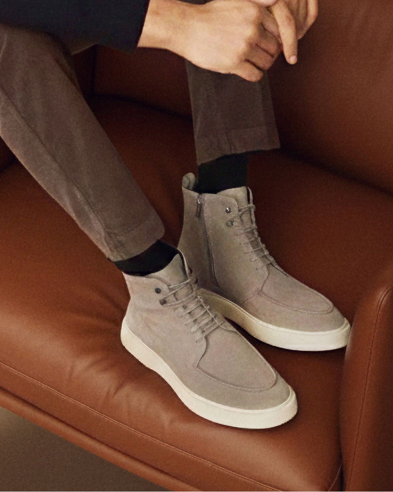 Beine eines Mannes mit hellgrauen Winterboots im Mokassin-Stil mit Schnürung und Reißverschluss, sitzend auf braunem Lederstuhl. Beine eines Mannes mit hellgrauen Winterboots im Mokassin-Stil mit Schnürung und Reißverschluss, sitzend auf braunem Lederstuhl.
