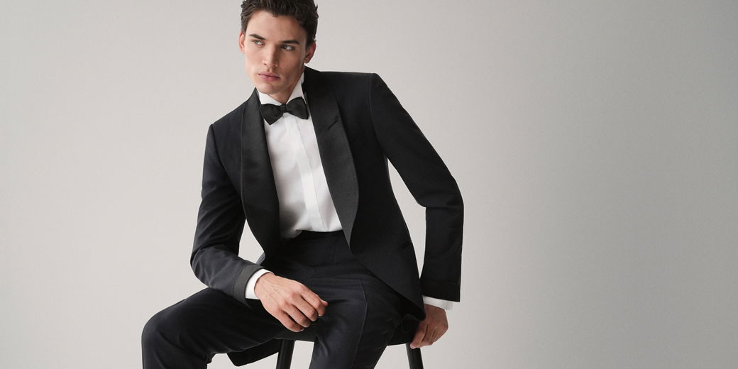 Junger Mann sitzt auf Hocker in elegantem schwarzen Smoking mit weißem Hemd und schwarzer Fliege vor schlichtem grauem Hintergrund.