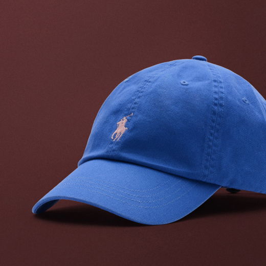 Blaue Baseball-Cap mit weißem Polo-Spieler-Logo, vor bordeauxrotem Hintergrund, seitlich fotografiert.