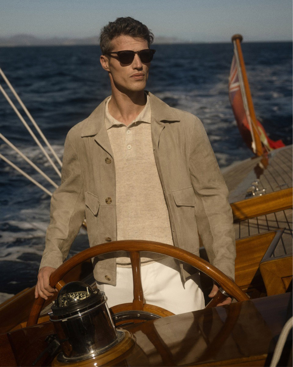 Mann mit dunkler Sonnenbrille steuert Segelboot, trägt weiße Hose, beiges Strickpolo und Velourslederjacke, Meer im Hintergrund.