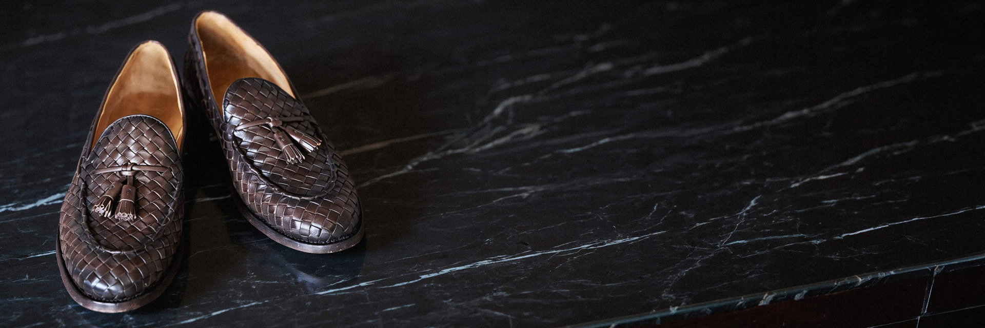 Braune Tasselloafer aus geflochtenem Leder mit Quasten auf marmoriertem, dunklem Untergrund liegend. Braune Tasselloafer aus geflochtenem Leder mit Quasten auf marmoriertem, dunklem Untergrund liegend.