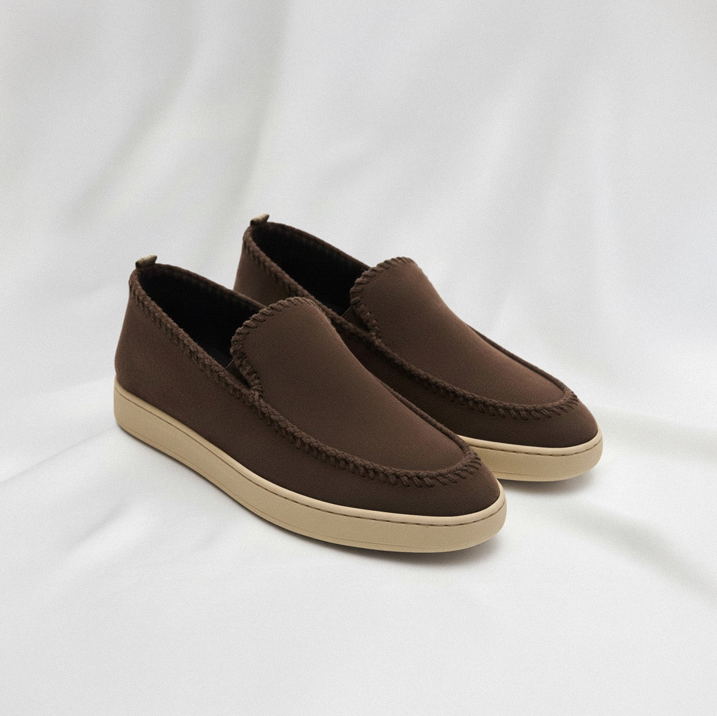 Braune Veloursleder Herren-Slipper mit beiger Gummisohle und Nahtdetails, auf hellgrauem Stoff liegend.