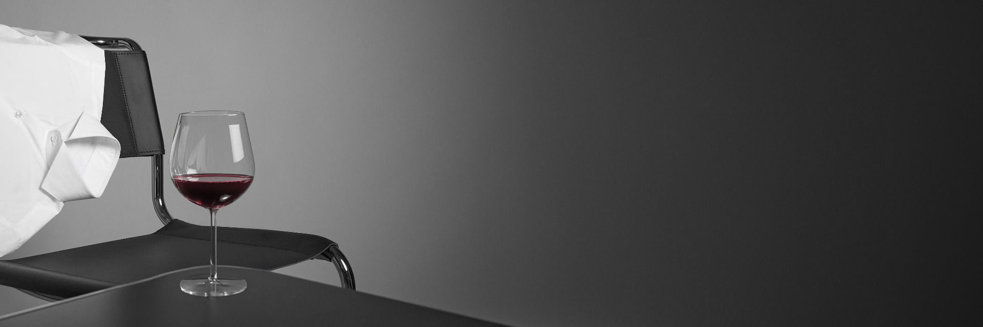 Weißes Businesshemd hängt über die Lehne eines schwarzen Stuhls, davor Glas mit Rotwein auf schwarzem Tisch. Weißes Businesshemd hängt über die Lehne eines schwarzen Stuhls, davor Glas mit Rotwein auf schwarzem Tisch.