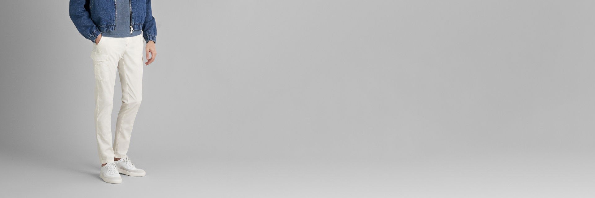 Männliches Model trägt offwhite Cargohose, weiße Sneaker und blaue Jeansjacke vor grauem Hintergrund. Männliches Model trägt offwhite Cargohose, weiße Sneaker und blaue Jeansjacke vor grauem Hintergrund.