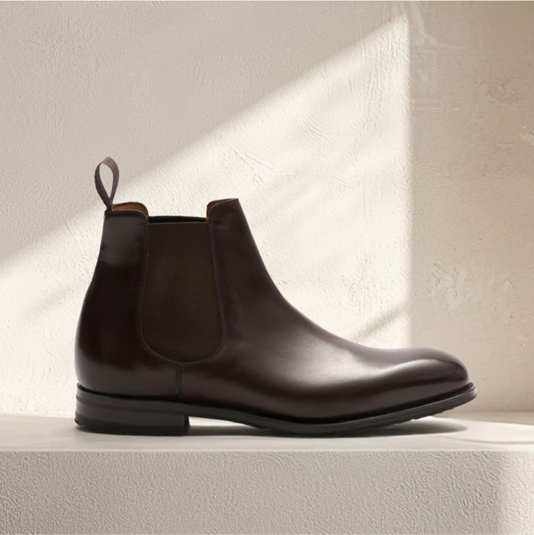 Dunkelbrauner Herren Chelsea-Boot aus Glattleder mit elastischem Einsatz und Zuglasche, vor weißer strukturierter Wand. Dunkelbrauner Herren Chelsea-Boot aus Glattleder mit elastischem Einsatz und Zuglasche, vor weißer strukturierter Wand.