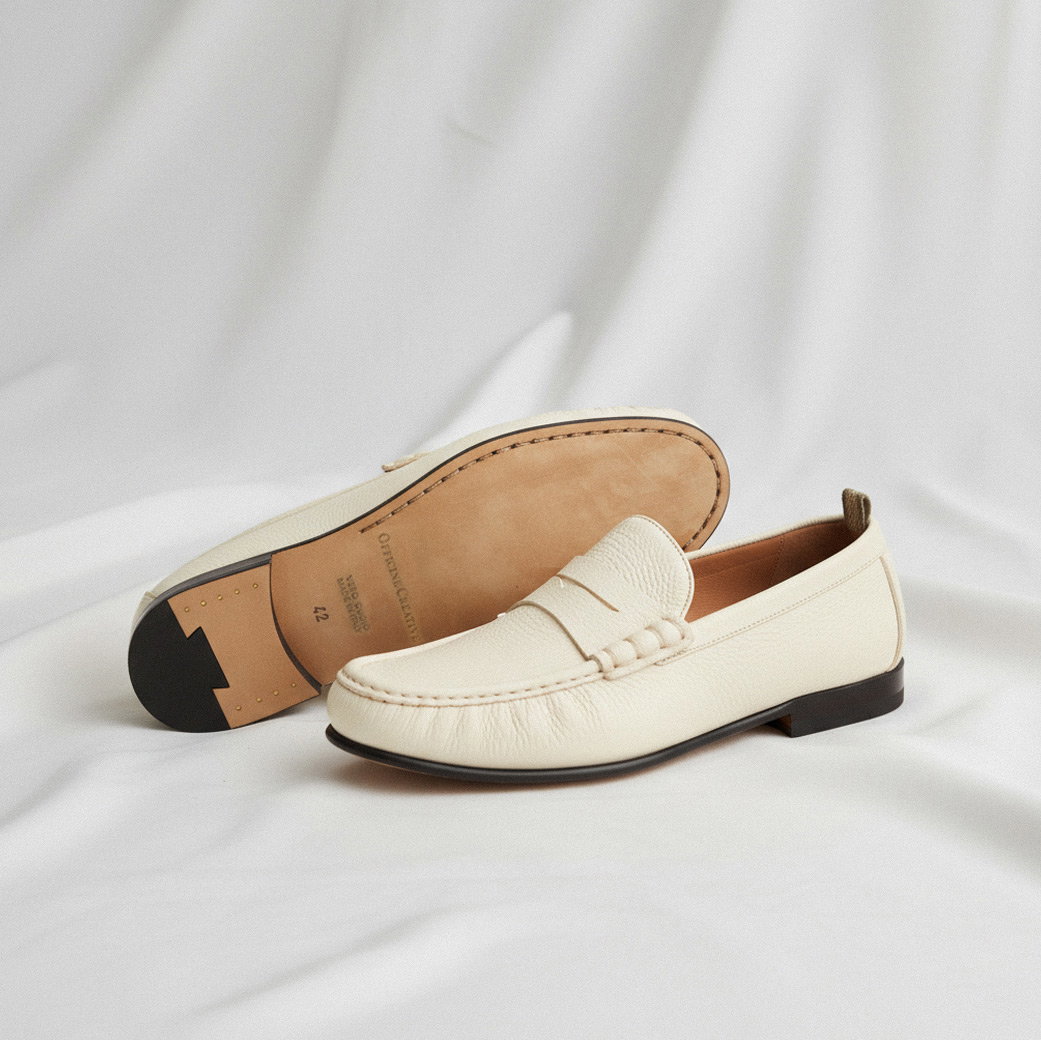 Cremefarbene Leder-Loafer mit dunkler Sohle, Ziernaht und dezenter Verzierung, auf hellem Stoff liegend.