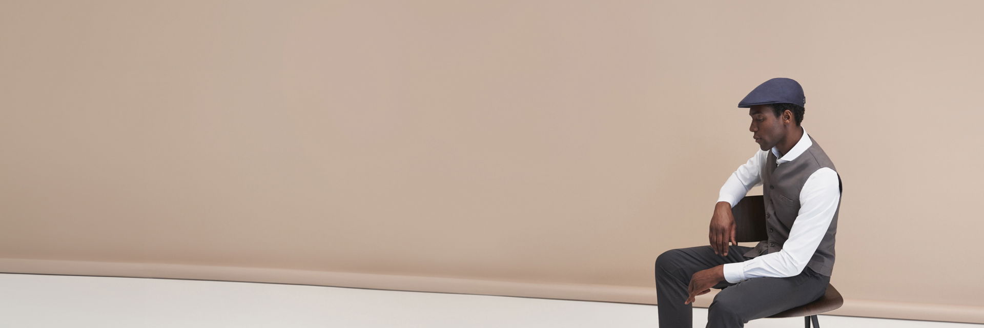 Junger Mann sitzt seitlich auf Stuhl, trägt weißes Hemd, graue Weste und Hose, blaue Schiebermütze vor beigem Hintergrund. Junger Mann sitzt seitlich auf Stuhl, trägt weißes Hemd, graue Weste und Hose, blaue Schiebermütze vor beigem Hintergrund.