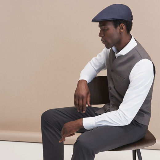 Mann sitzt seitlich auf Stuhl, trägt weiße Hemd, graue Weste, dunkelgraue Hose und dunkelblaue Schiebermütze vor beigem Hintergrund.